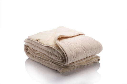 Reversible blanket with one side in soft cream organic cotton quilted with wool padding and the other side in natural beige merino wool, shown folded to highlight both textures. Made by Woolland