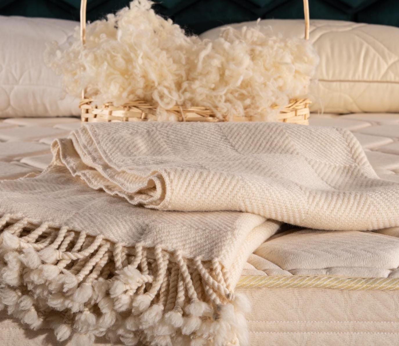 Folded merino wool blanket with tassels on a bed with a basket of raw wool yarn. Handcrafted by Woolland