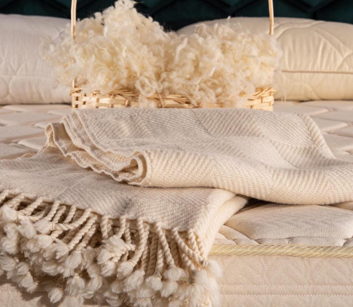 Folded merino wool blanket with tassels on a bed with a basket of raw wool yarn. Handcrafted by Woolland