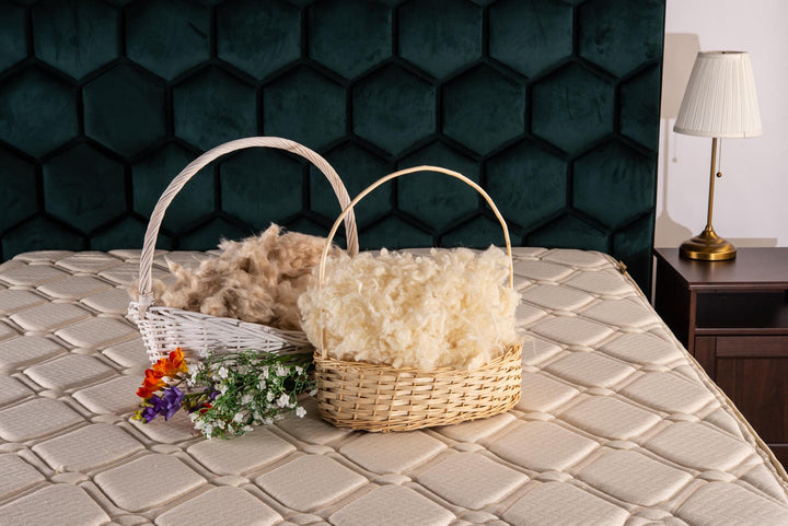 Two woven baskets filled with raw wool and kapok fibers on a textured bed with a dark green hexagonal wall and a lamp in the background.
