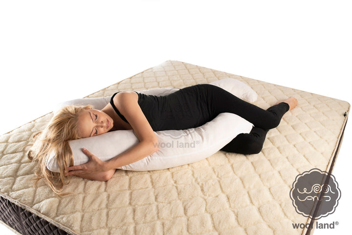A pregnant woman is laying on a bed snuggling a white organic fibers u-shaped pregnancy pillow handmade by Woolland