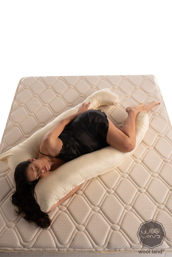 A pregnant lady is laying on a bed snuggling a natural wool and cotton u-shaped pregnancy maternity pillow handcrafted by Woolland