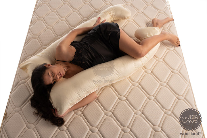 A beautiful pregnant woman laying on a bed snuggling a cotton sateen and raw wool u-shaped pregnancy pillow handcrafted by Woolland