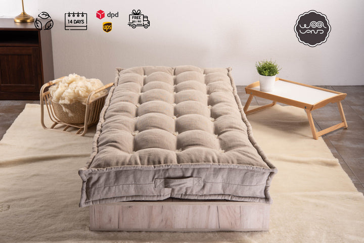 A mattress topped with merino wool and linen, displayed on a bed frame with a linen cover, set against a neutral background with a wicker chair and a small wooden side table in the background.