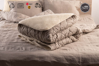 A folded natural linen and wool blanket on a bed, with a pillow and a decorative item on top.