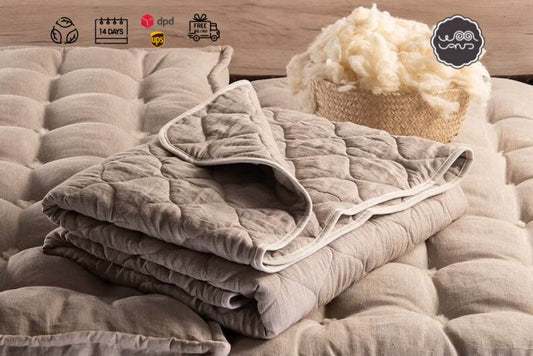 A folded natural linen quilt with a woolen filling, displayed on a bed with a plush toy on the side, and a label indicating the product's certification.
